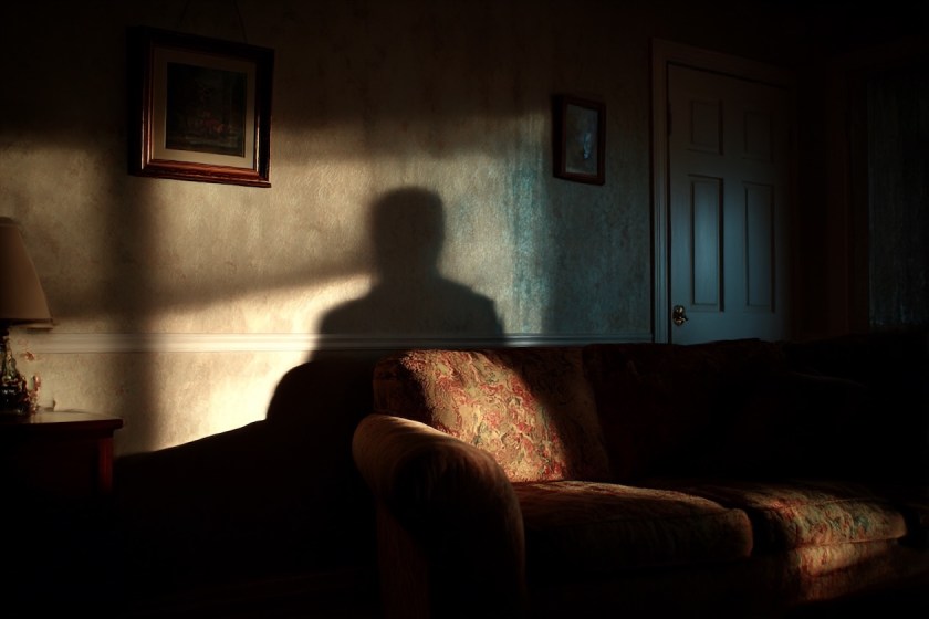 Surreal illustration of a living room where the light hides behind a sofa while an oversized shadow stretches across the floor