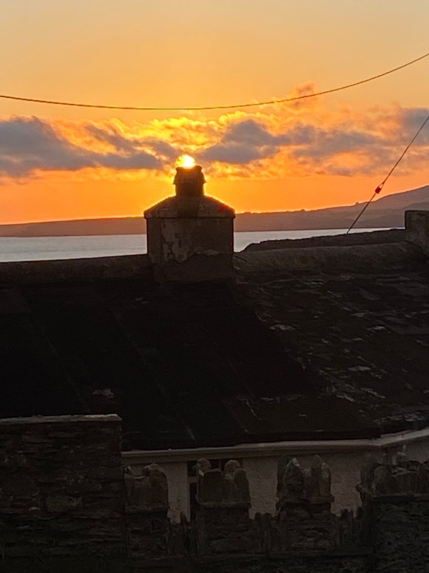 A stone rooftop and chimney stack silhouetted against a vivid orange sunset on the west coast of Ireland, with a calm sea and distant headland visible on the horizon. A telegraph wire crosses the sky diagonally in the upper portion of the image.